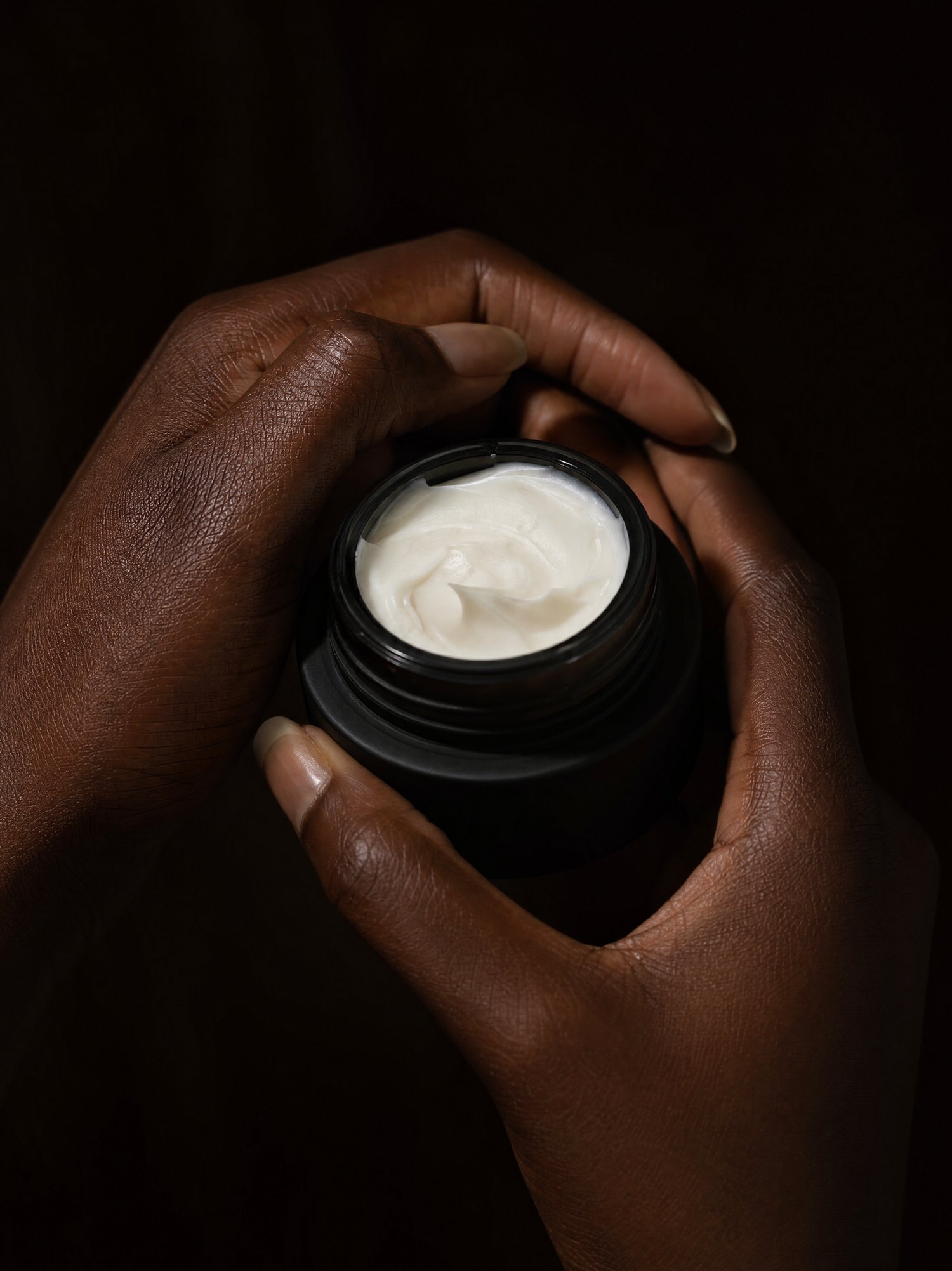 Hands cradling an open matte-black jar of MelaSil cream in warm low light